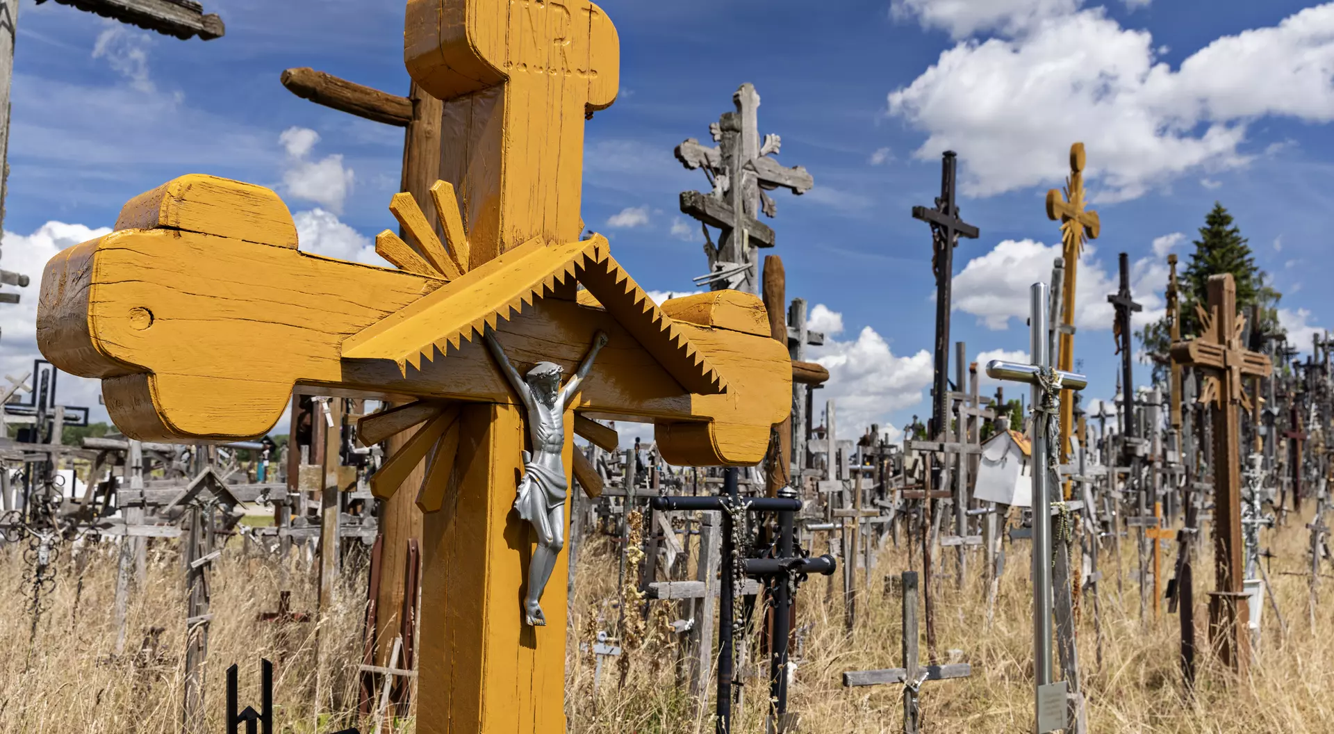 A wooden cross with a figure of Christ in the foreground, surrounded by numerous other crosses in a grassy cemetery.
