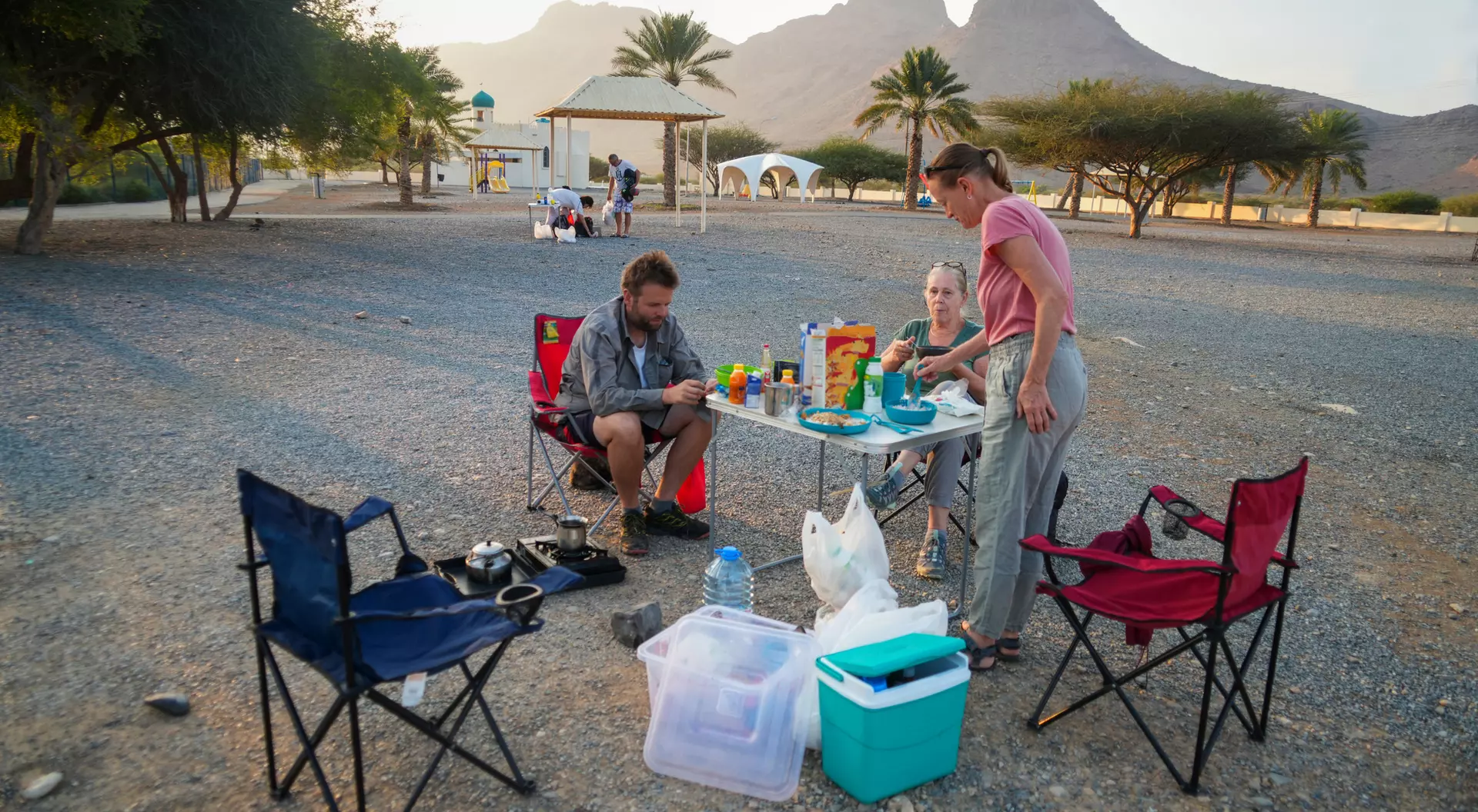 Two people prepare food at a campsite with mountains in the background, surrounded by empty chairs and camping gear.