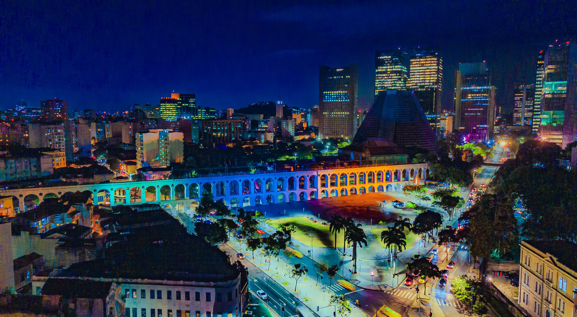 A nighttime aerial view of a vibrant cityscape with illuminated buildings and an arched aqueduct.