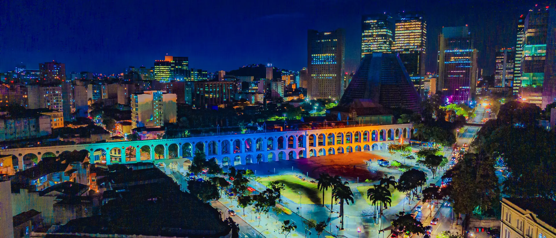 A nighttime aerial view of a vibrant cityscape with illuminated buildings and an arched aqueduct.