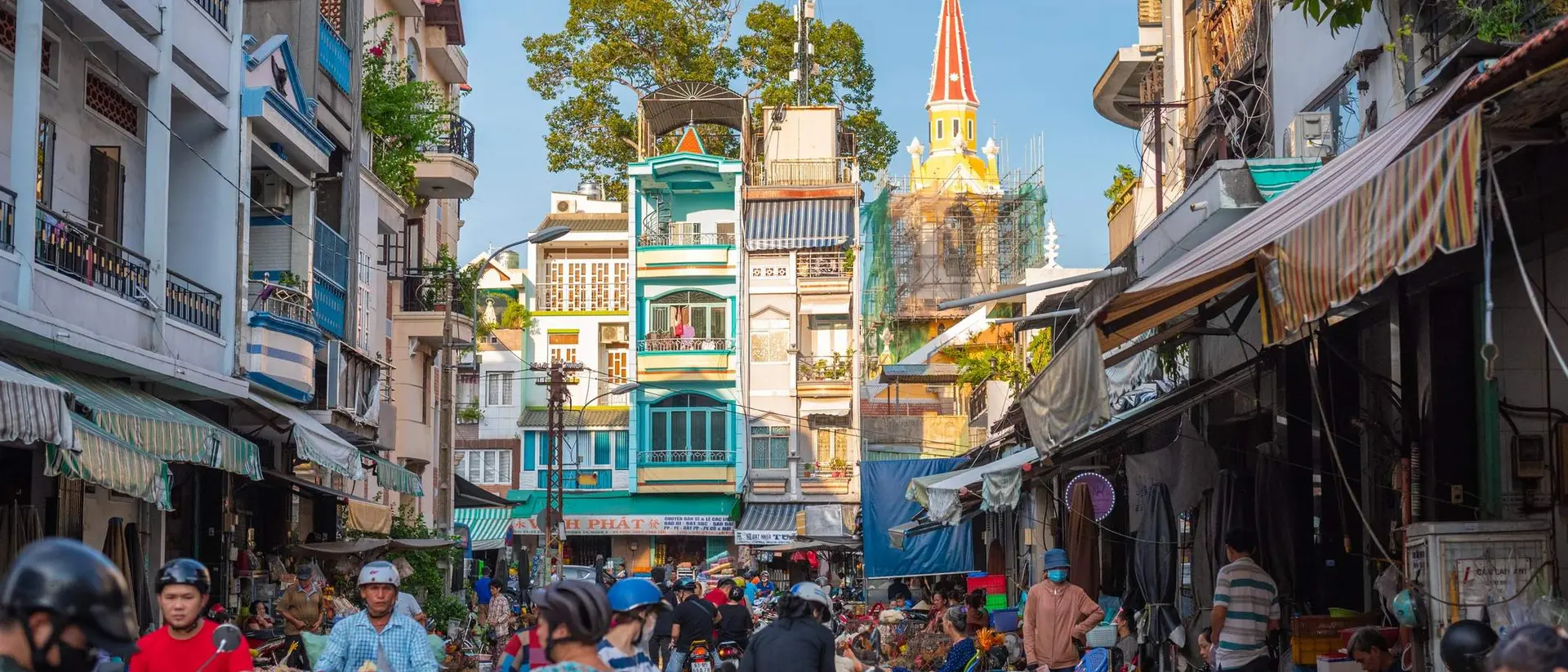 Bustling street scene filled with people and motorbikes, tall buildings, and a church steeple in the background.