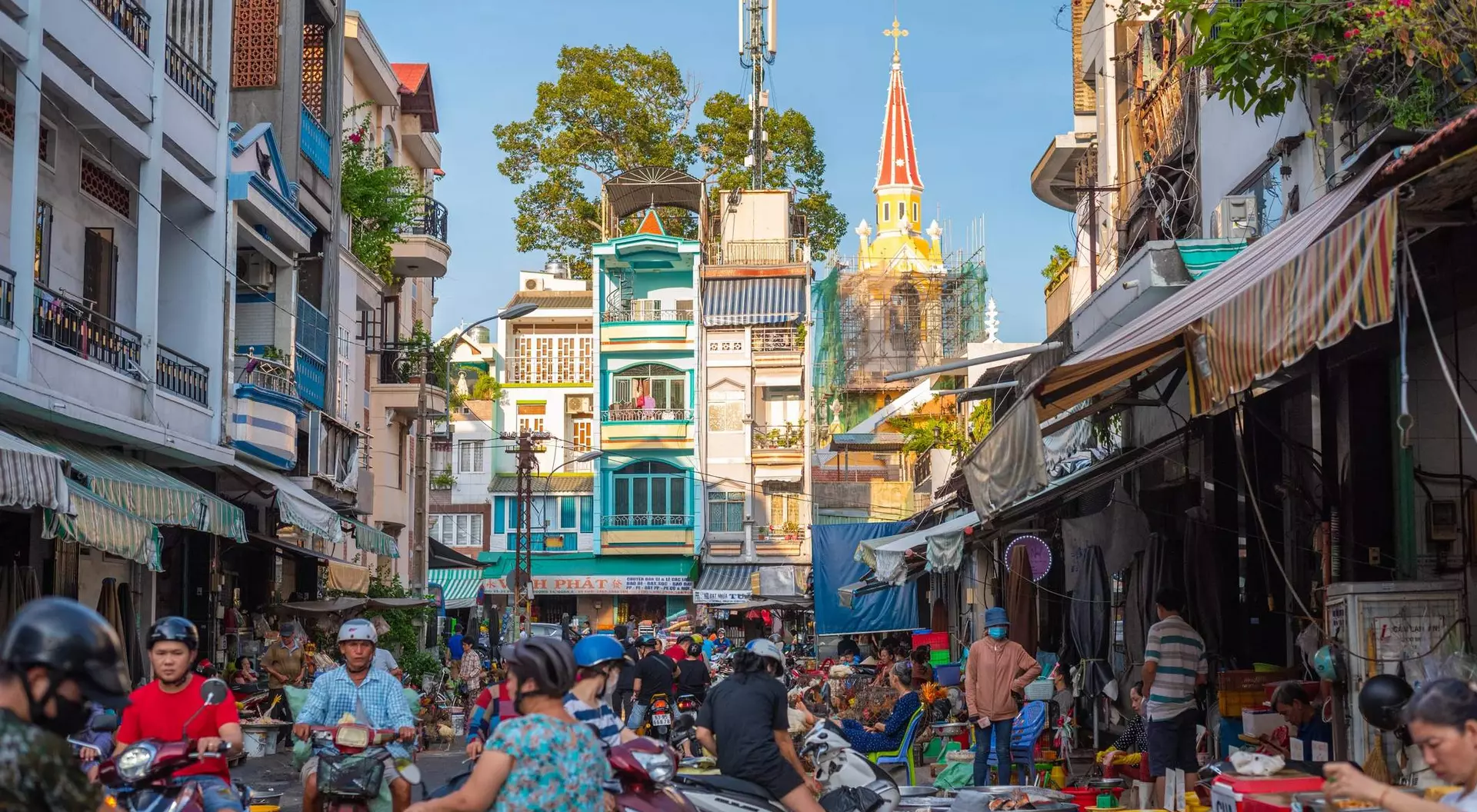 Bustling street scene filled with people and motorbikes, tall buildings, and a church steeple in the background.