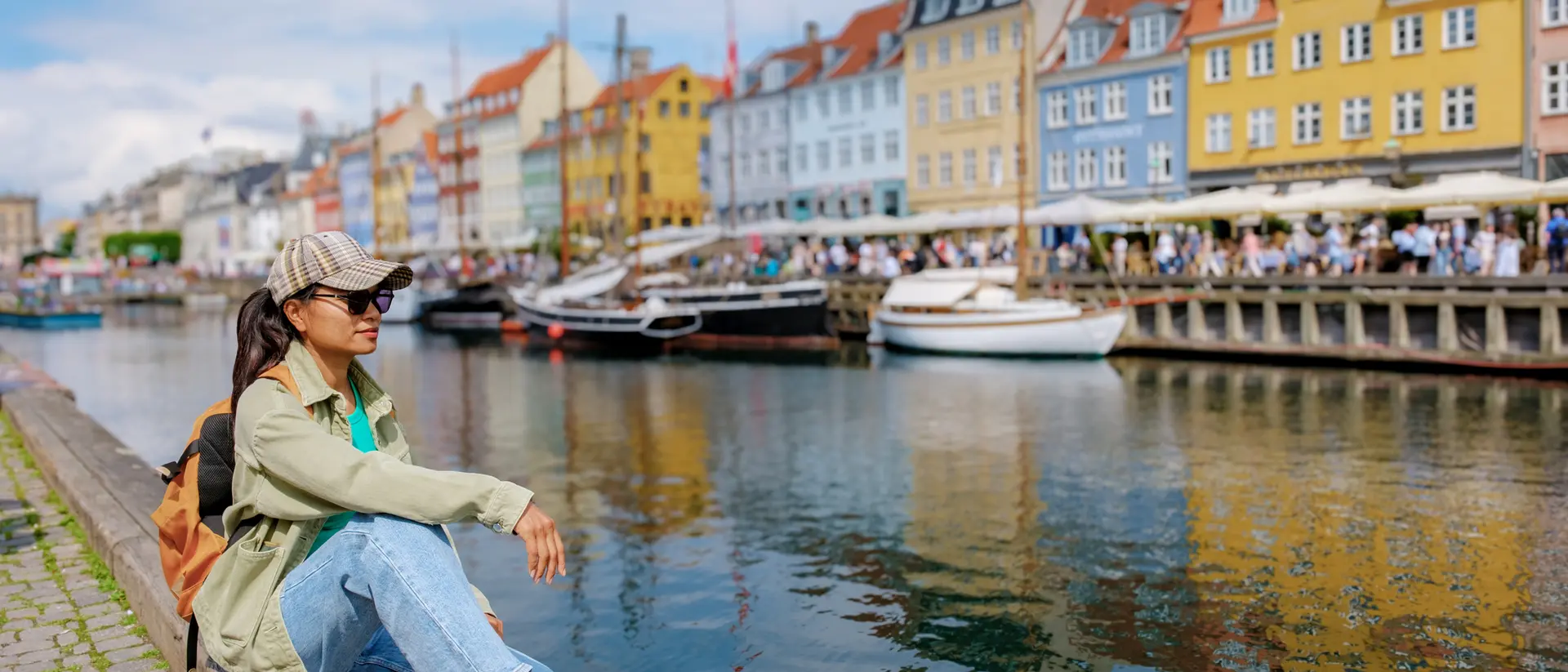 A person sits on a dock by a colorful waterfront, with boats and vibrant buildings reflecting in the water.
