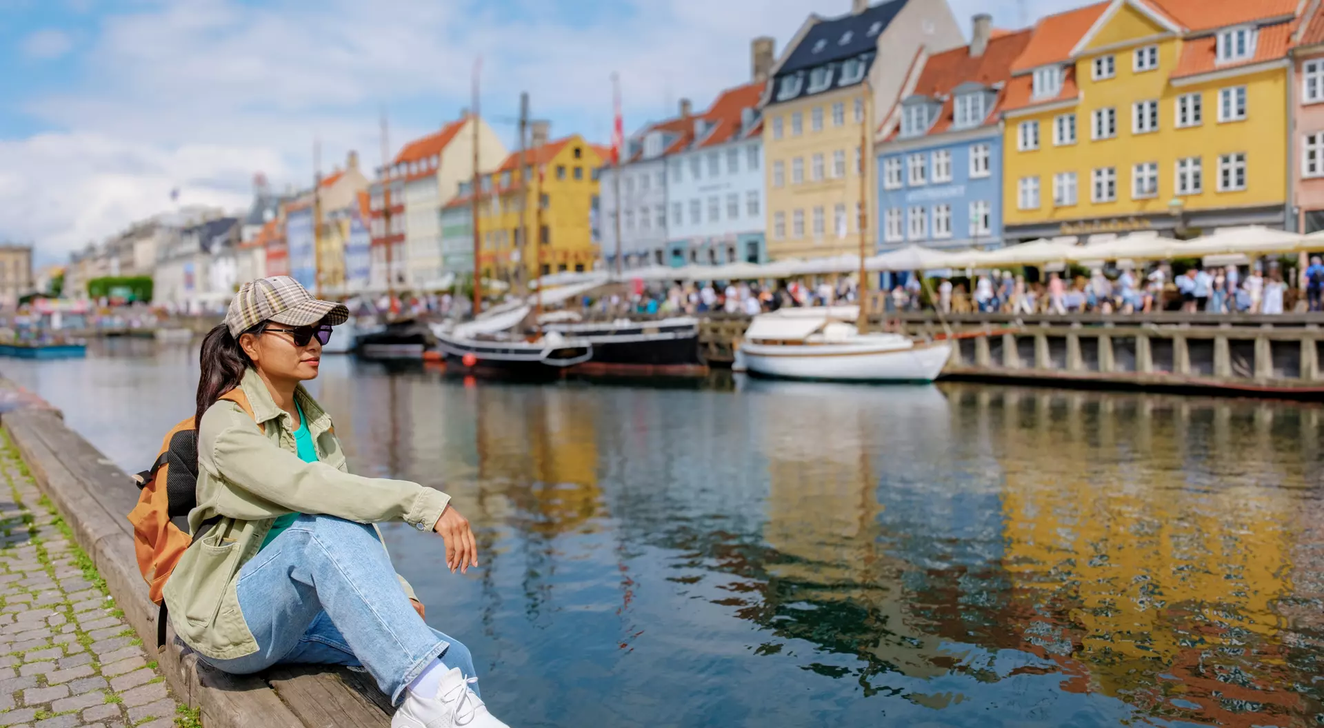 A person sits on a dock by a colorful waterfront, with boats and vibrant buildings reflecting in the water.