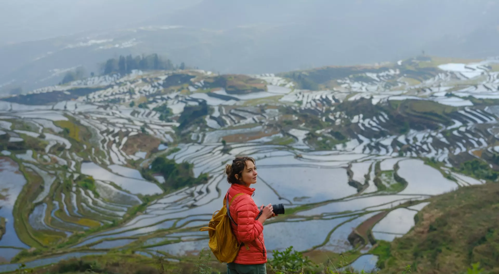 Person in a red jacket stands on a hillside, overlooking terraced rice fields reflecting water under a cloudy sky.
