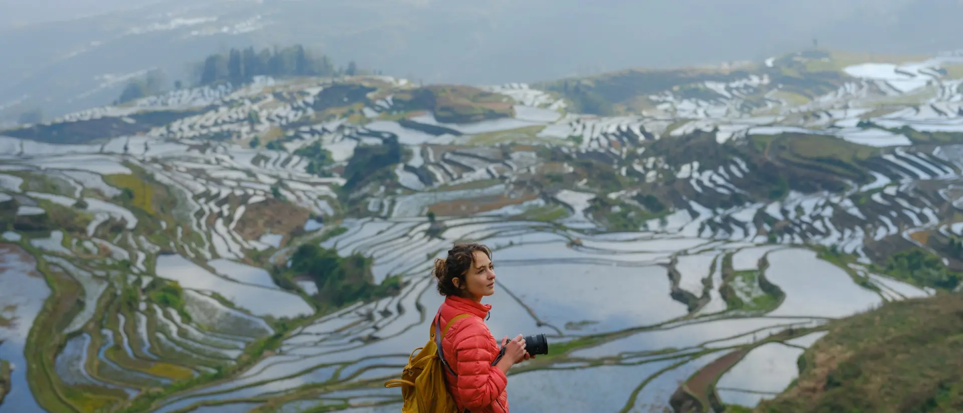 Person in a red jacket stands on a hillside, overlooking terraced rice fields reflecting water under a cloudy sky.
