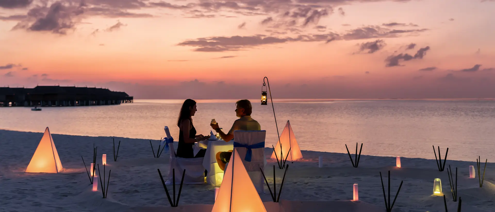 A couple enjoys a sunset dinner on the beach, surrounded by glowing lanterns and soft sand.