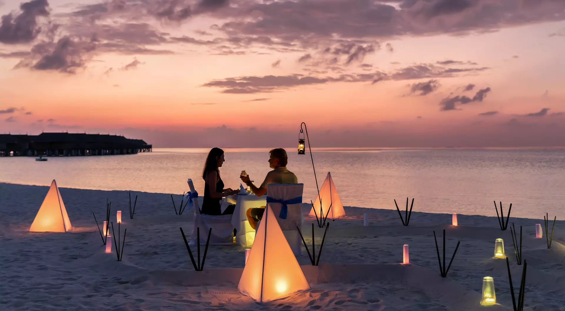 A couple enjoys a sunset dinner on the beach, surrounded by glowing lanterns and soft sand.