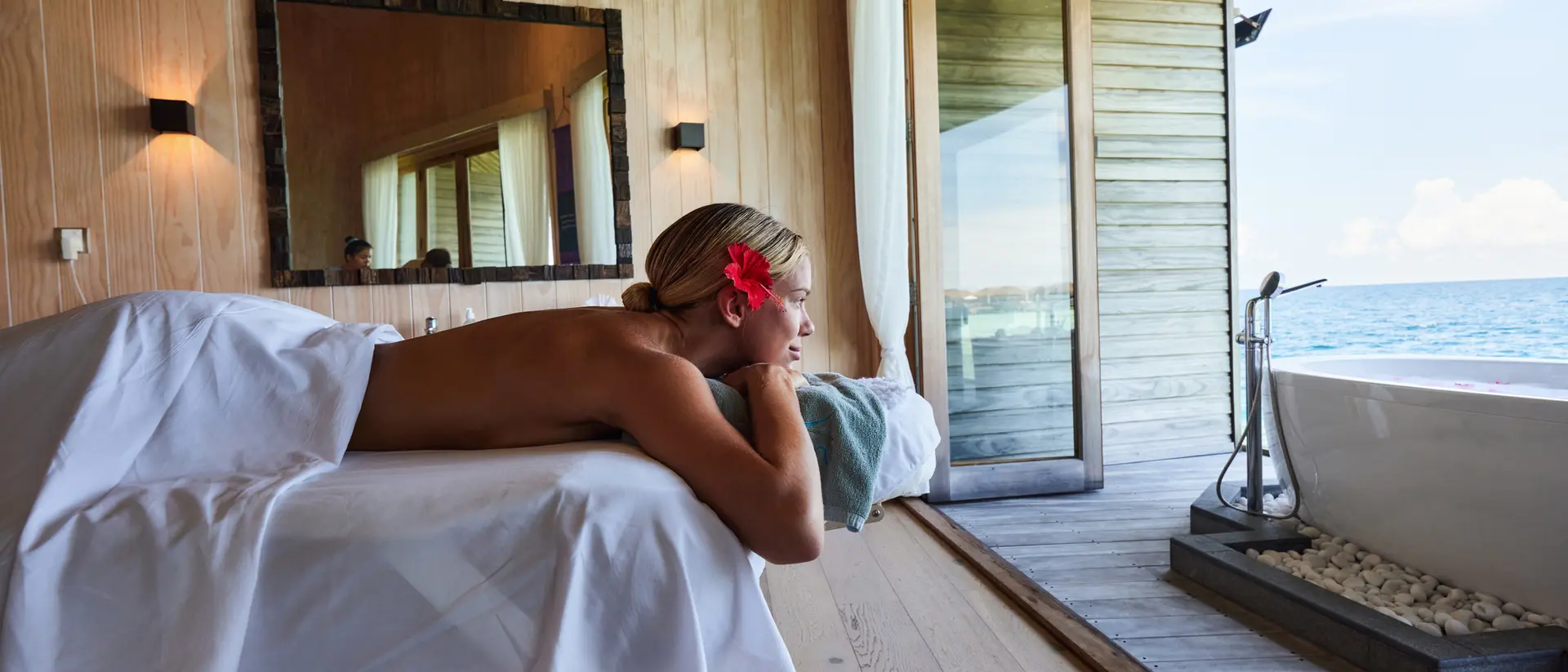 A person enjoys a relaxing massage in a serene spa with a view of the ocean, adorned with a flower in their hair.