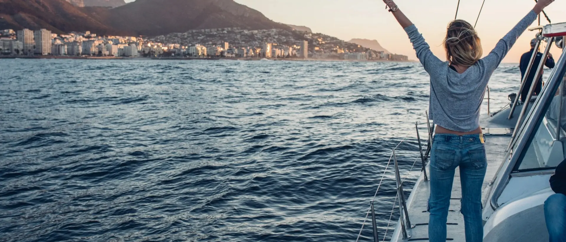 Person on a sailboat raises arms in joy, with mountains and a city skyline in the background at sunset.