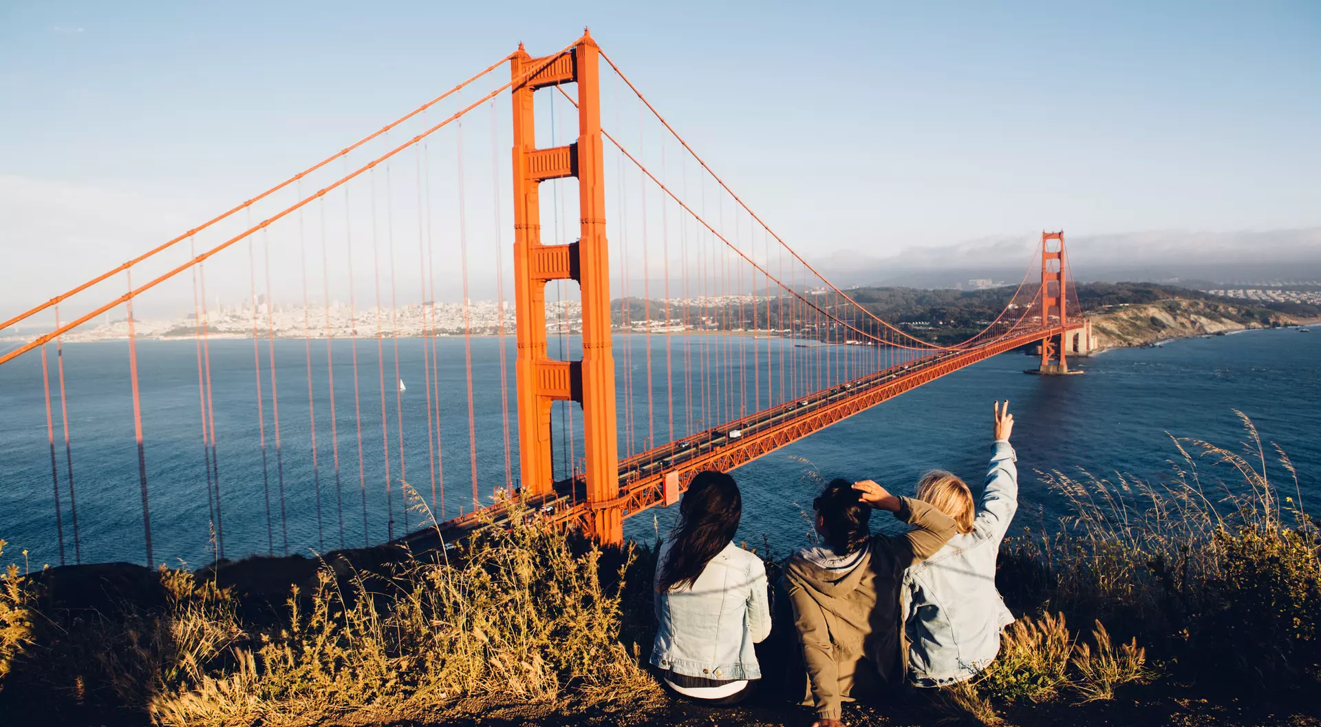 Three people sit on a hillside overlooking the Golden Gate Bridge, enjoying the view on a sunny day.