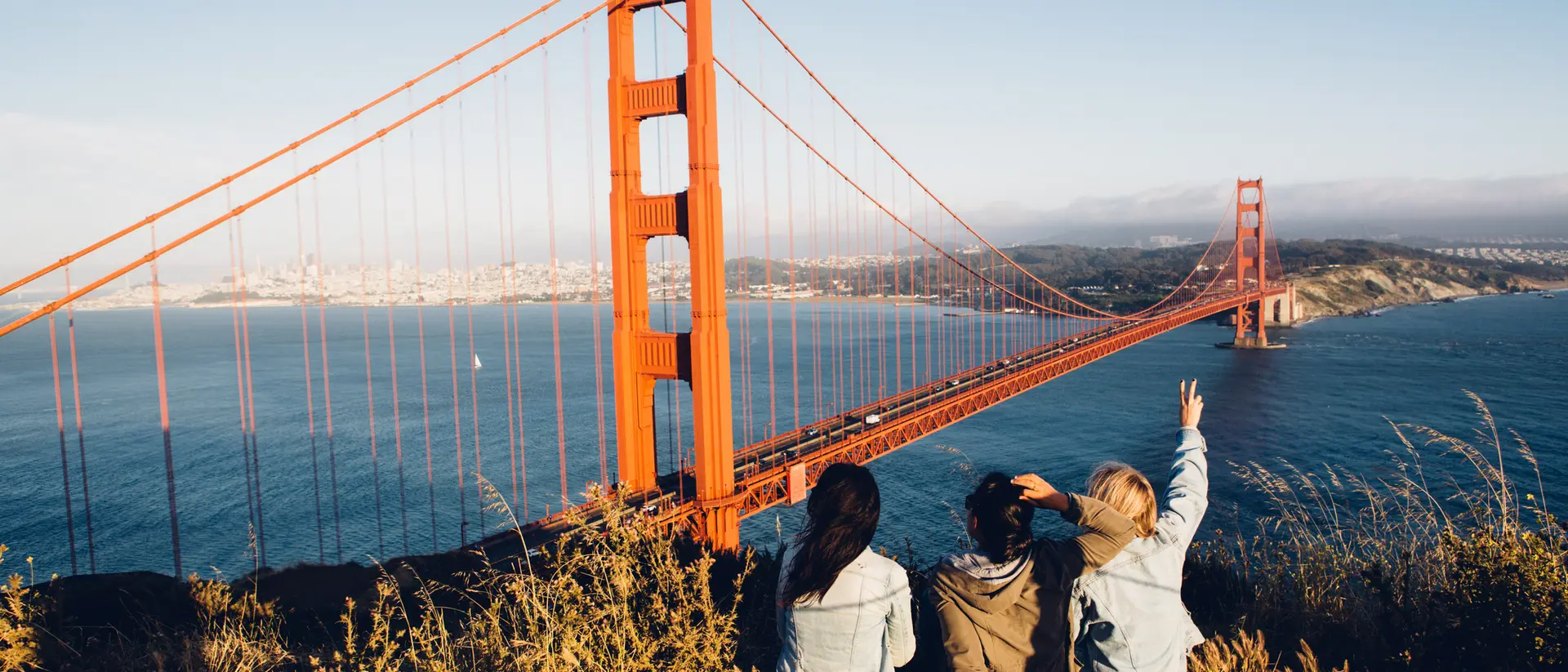 Three people sit on a hillside overlooking the Golden Gate Bridge, enjoying the view on a sunny day.