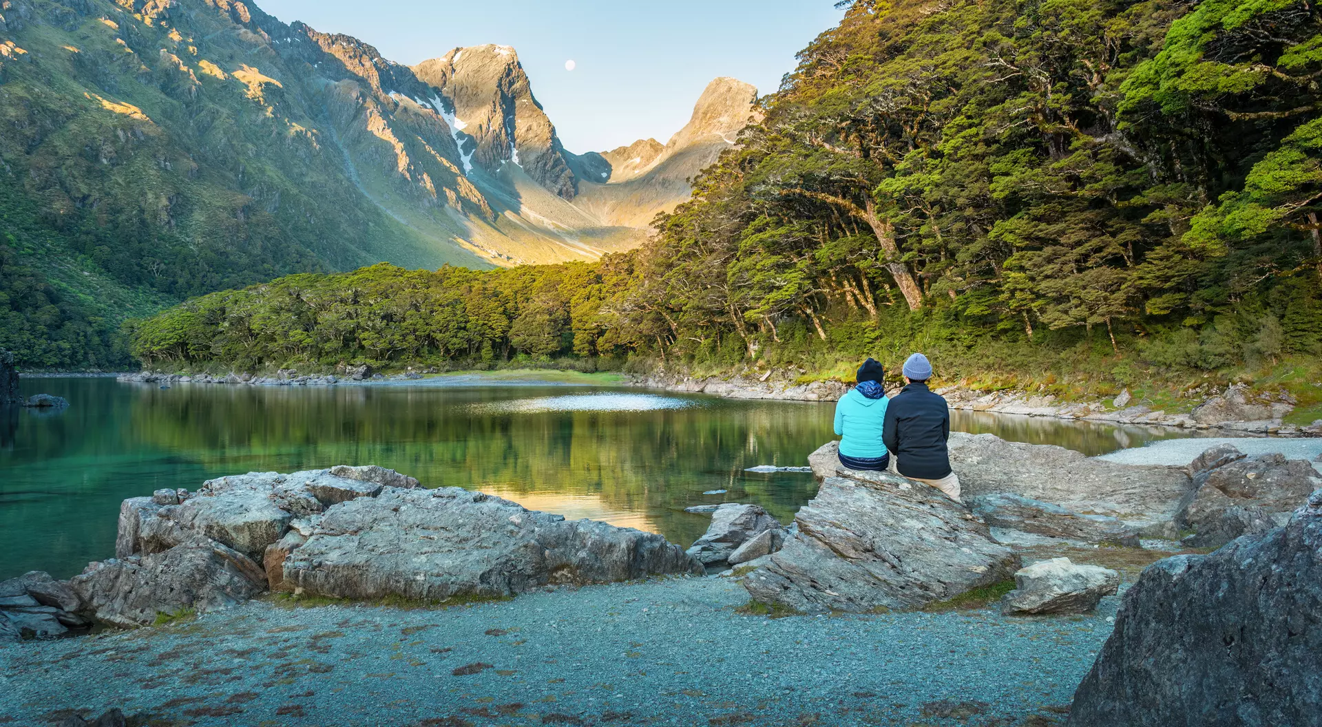 Two people sit on a rock by a tranquil lake, surrounded by mountains and lush greenery during a serene morning.