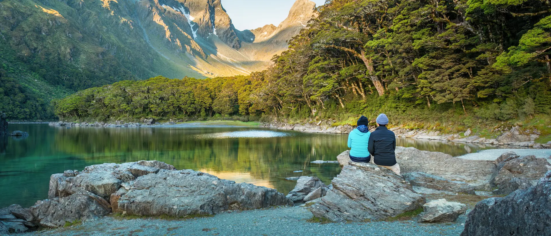 Two people sit on a rock by a tranquil lake, surrounded by mountains and lush greenery during a serene morning.