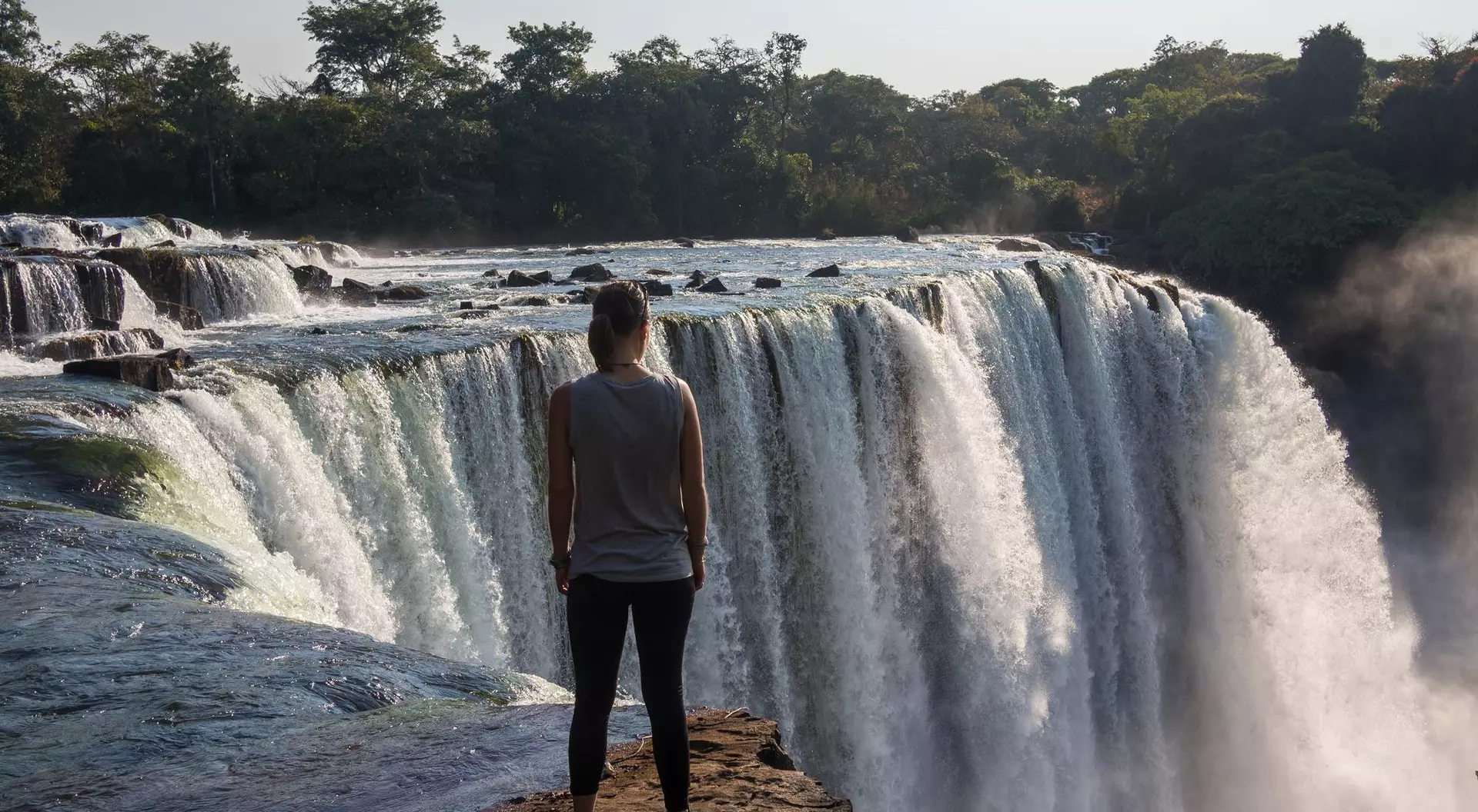 A person stands on the edge of a rocky cliff, looking out at a large waterfall surrounded by lush greenery.