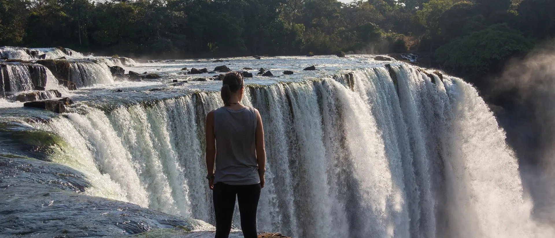 A person stands on the edge of a rocky cliff, looking out at a large waterfall surrounded by lush greenery.
