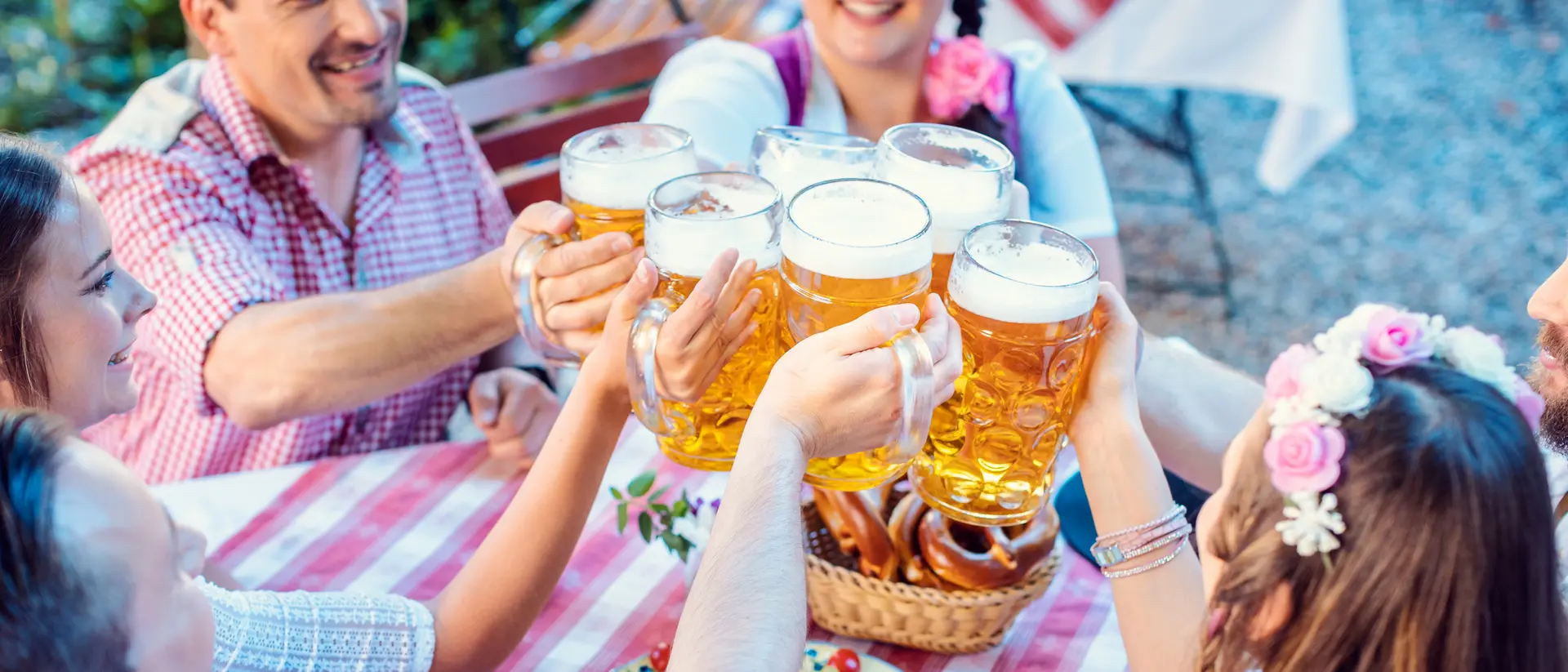 Group of people cheerfully toasting with large beers at a outdoor table, surrounded by pretzels and a festive atmosphere.