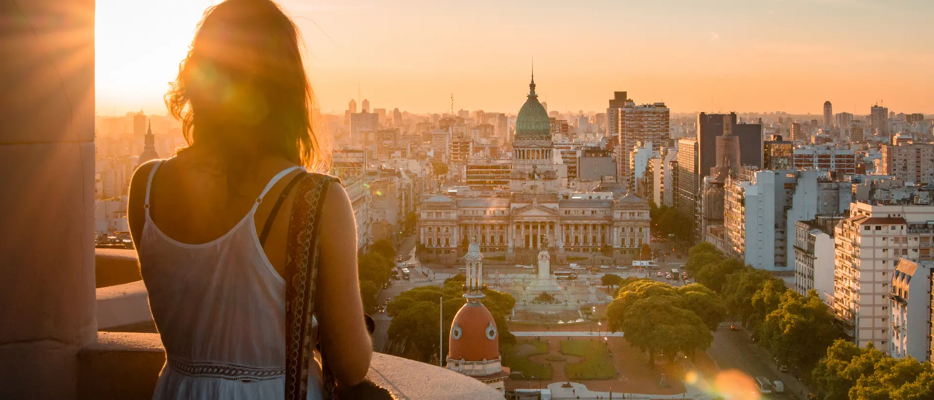 Silhouette of a person overlooking a city at sunset, with buildings and a central plaza visible below.