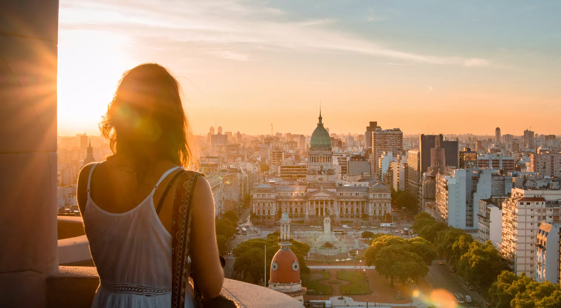 Silhouette of a person overlooking a city at sunset, with buildings and a central plaza visible below.