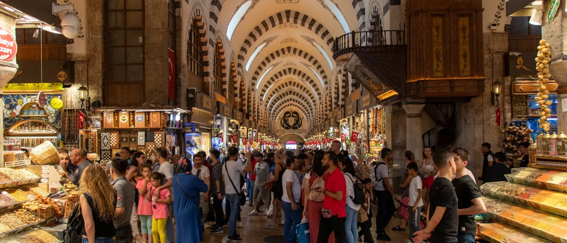 Busy market interior filled with shoppers, stalls of colorful goods, and a high, patterned ceiling.