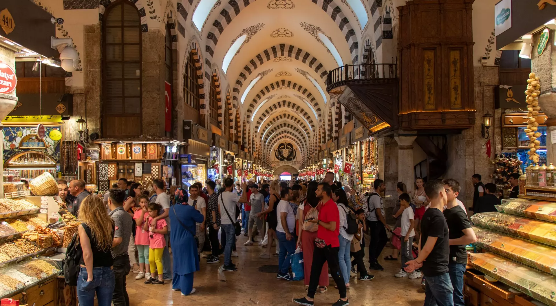 Busy market interior filled with shoppers, stalls of colorful goods, and a high, patterned ceiling.