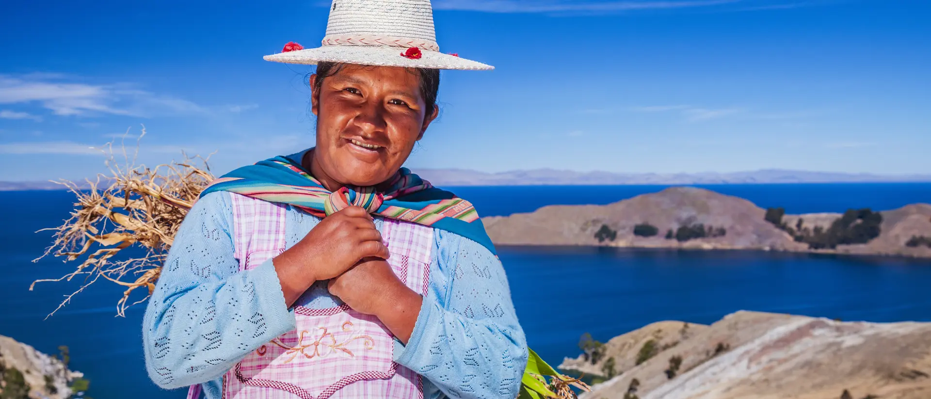 A smiling woman in traditional attire stands near a lake, holding plants, with mountains and a clear blue sky in the background.