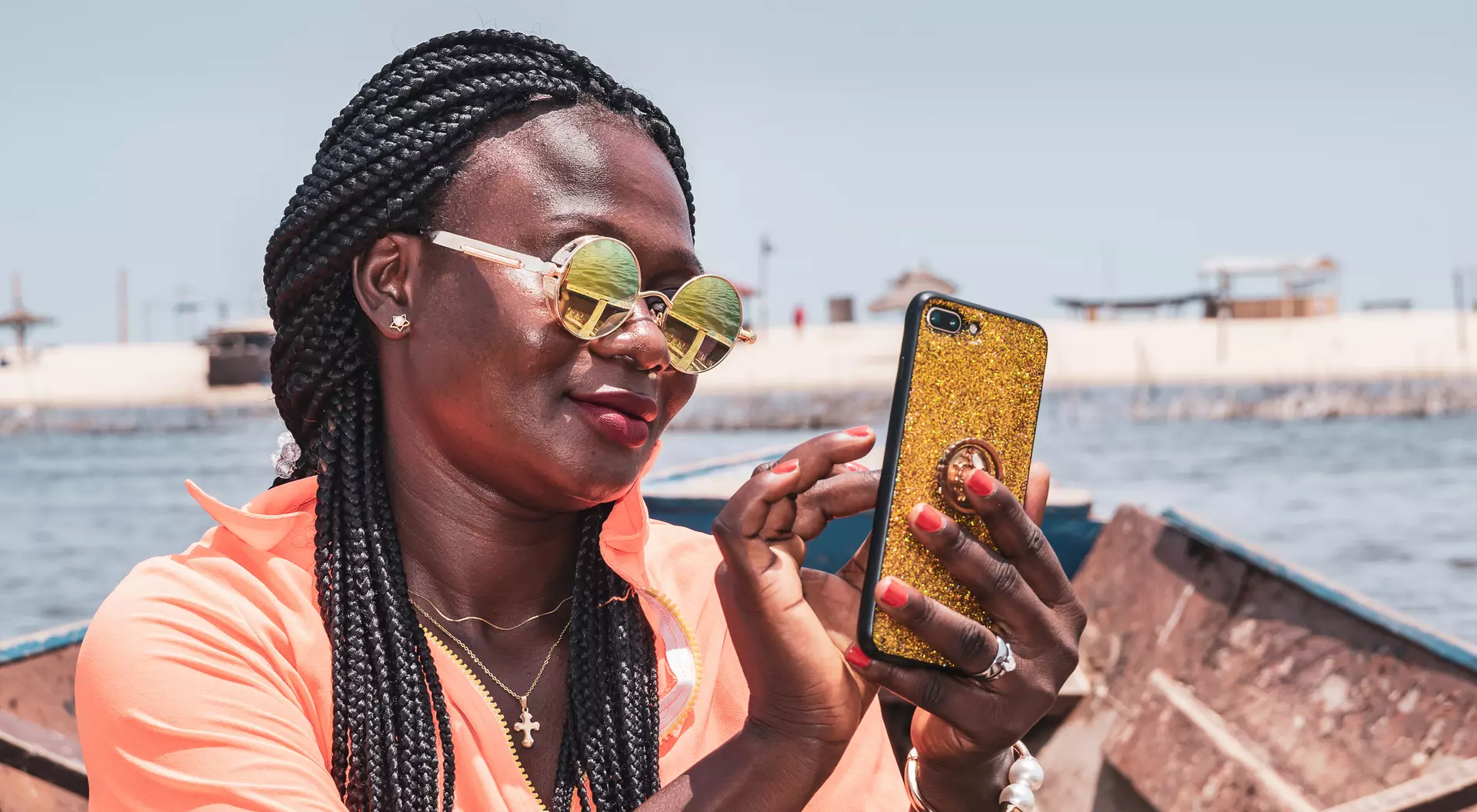 A woman with braided hair and sunglasses smiles while using a phone on a boat near the shore.