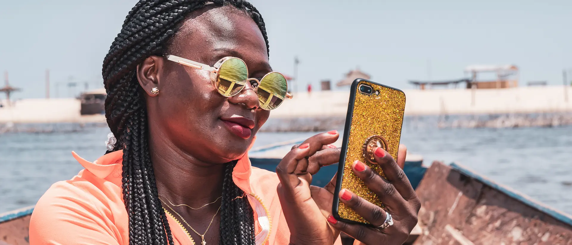 A woman with braided hair and sunglasses smiles while using a phone on a boat near the shore.