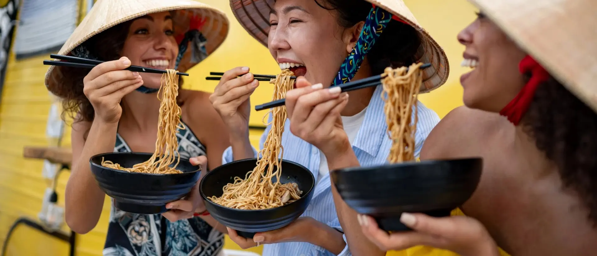 Three women in traditional hats enjoy noodles from bowls, laughing together in front of a bright yellow food truck.