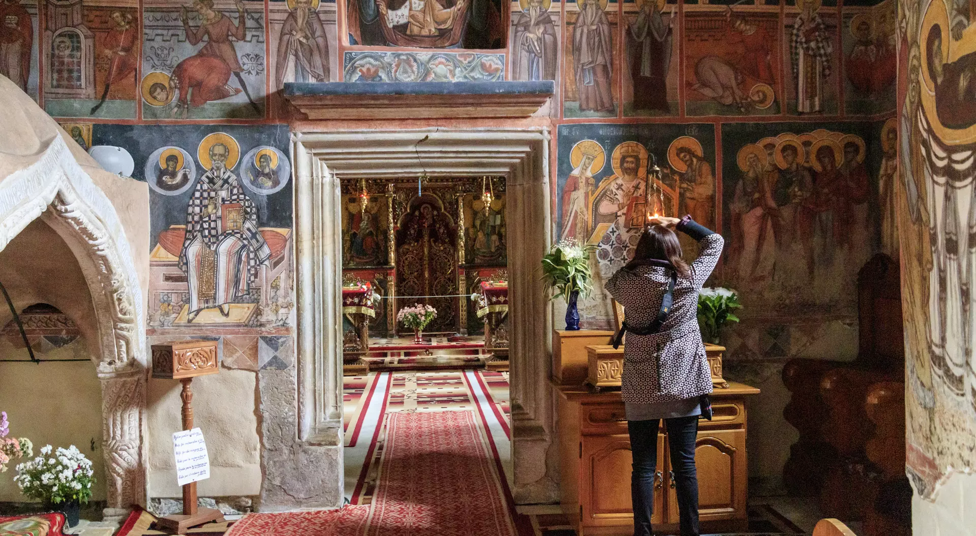 A person admires colorful frescoes inside a church, with floral arrangements and a dimly lit altar in view.