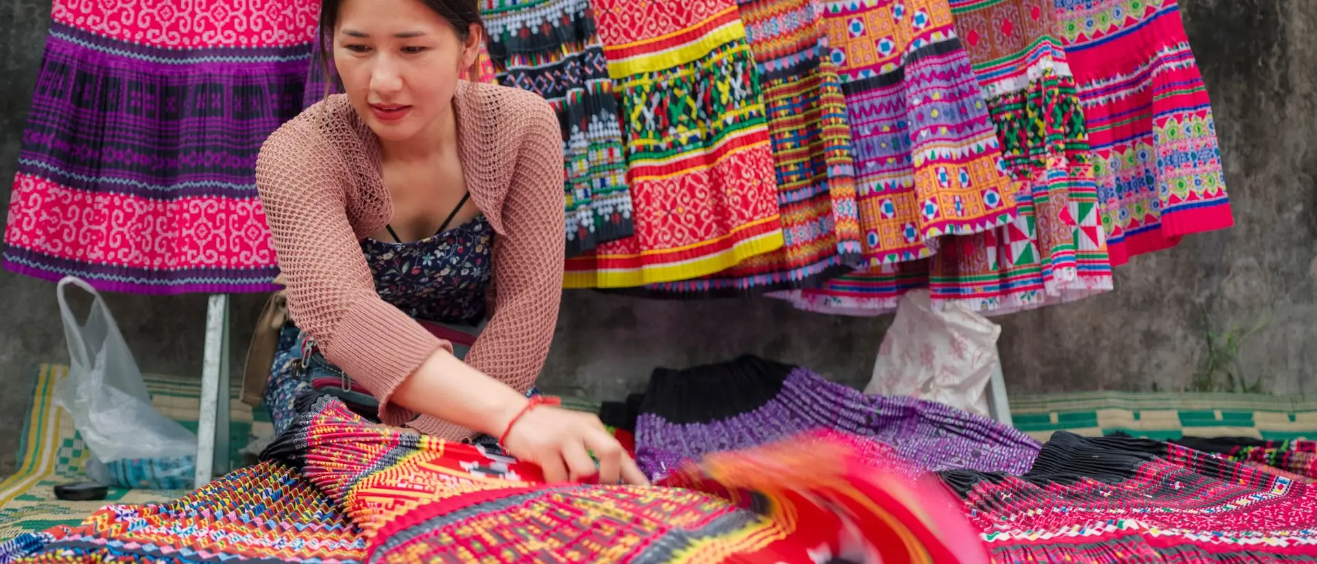 A woman arranges colorful, patterned textiles at a market, with vibrant traditional garments hanging in the background.