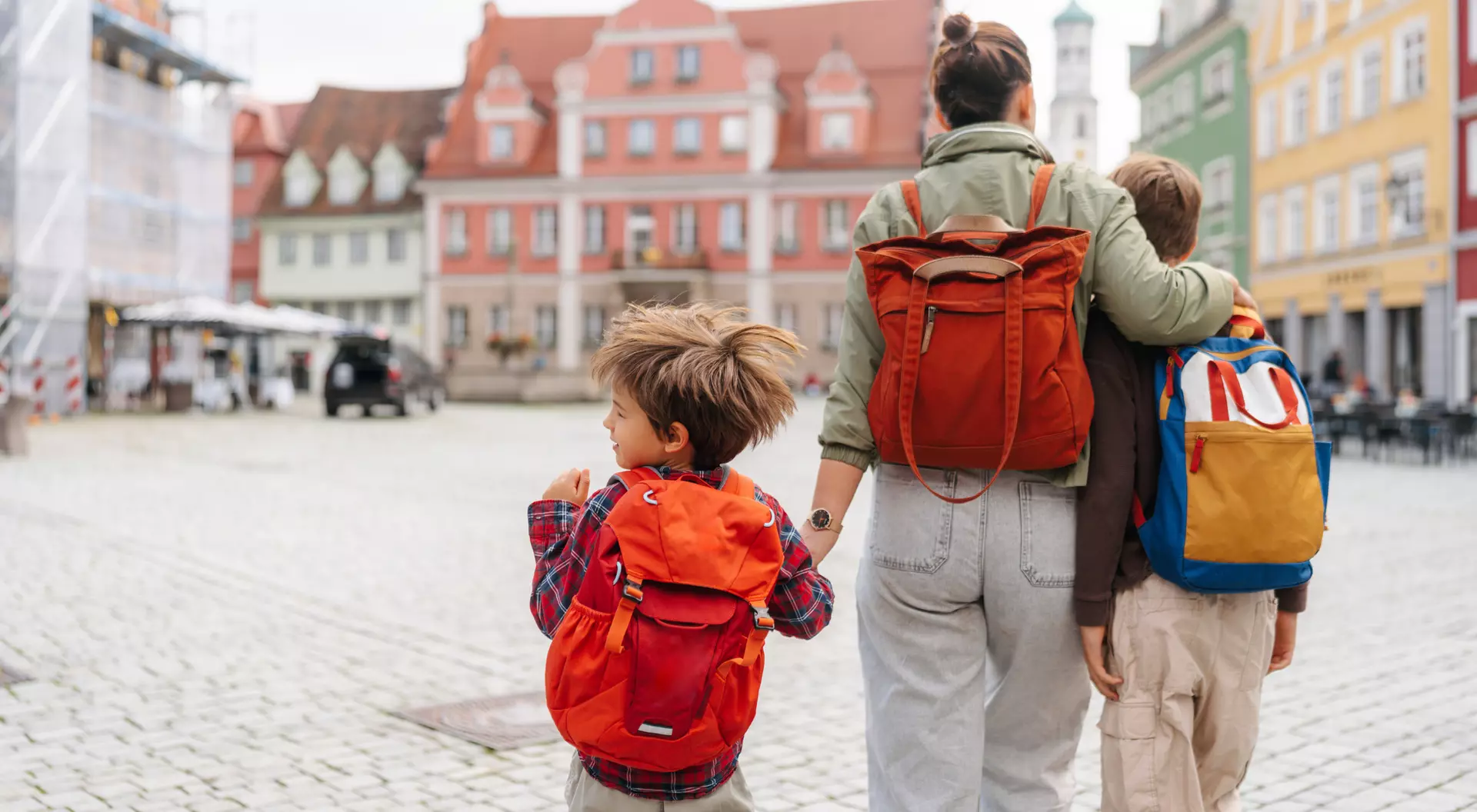 A woman and two children, all with backpacks, walk together in a cobblestone square with colorful buildings in the background.