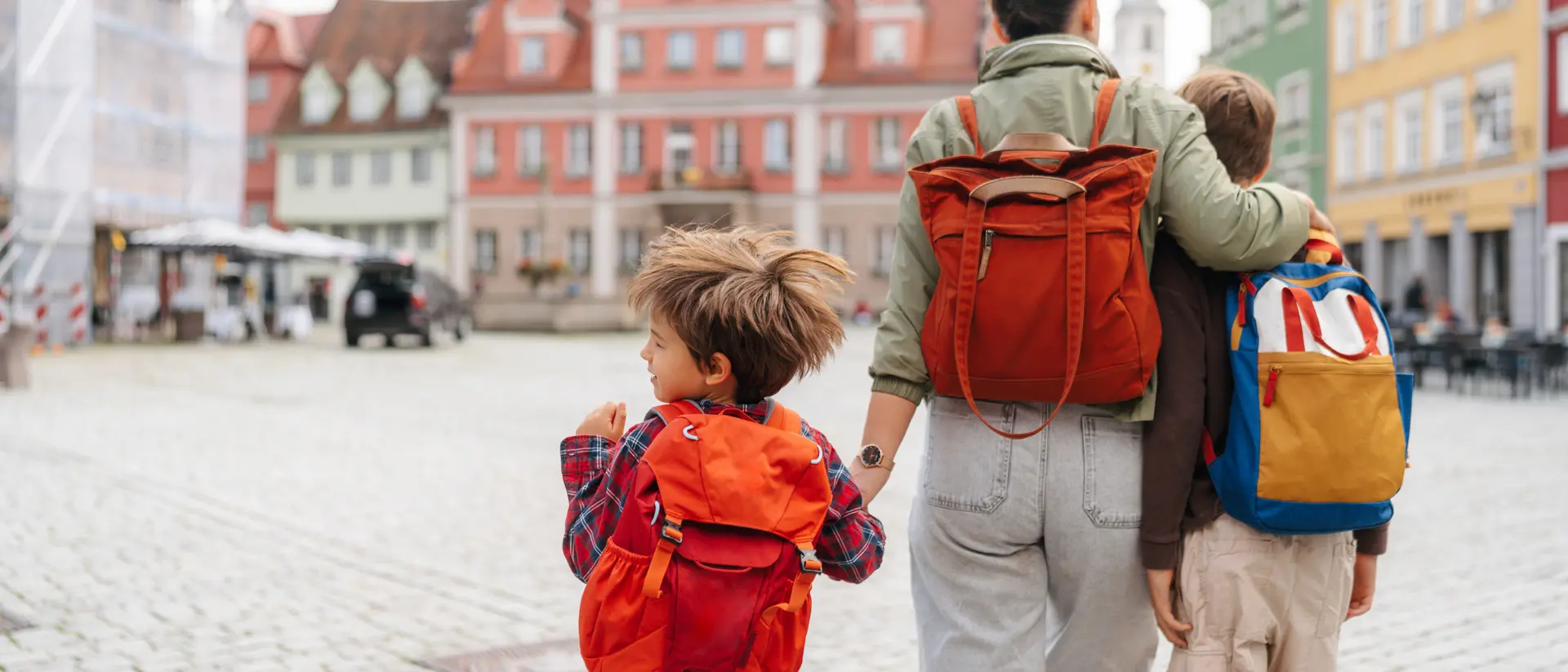 A woman and two children, all with backpacks, walk together in a cobblestone square with colorful buildings in the background.