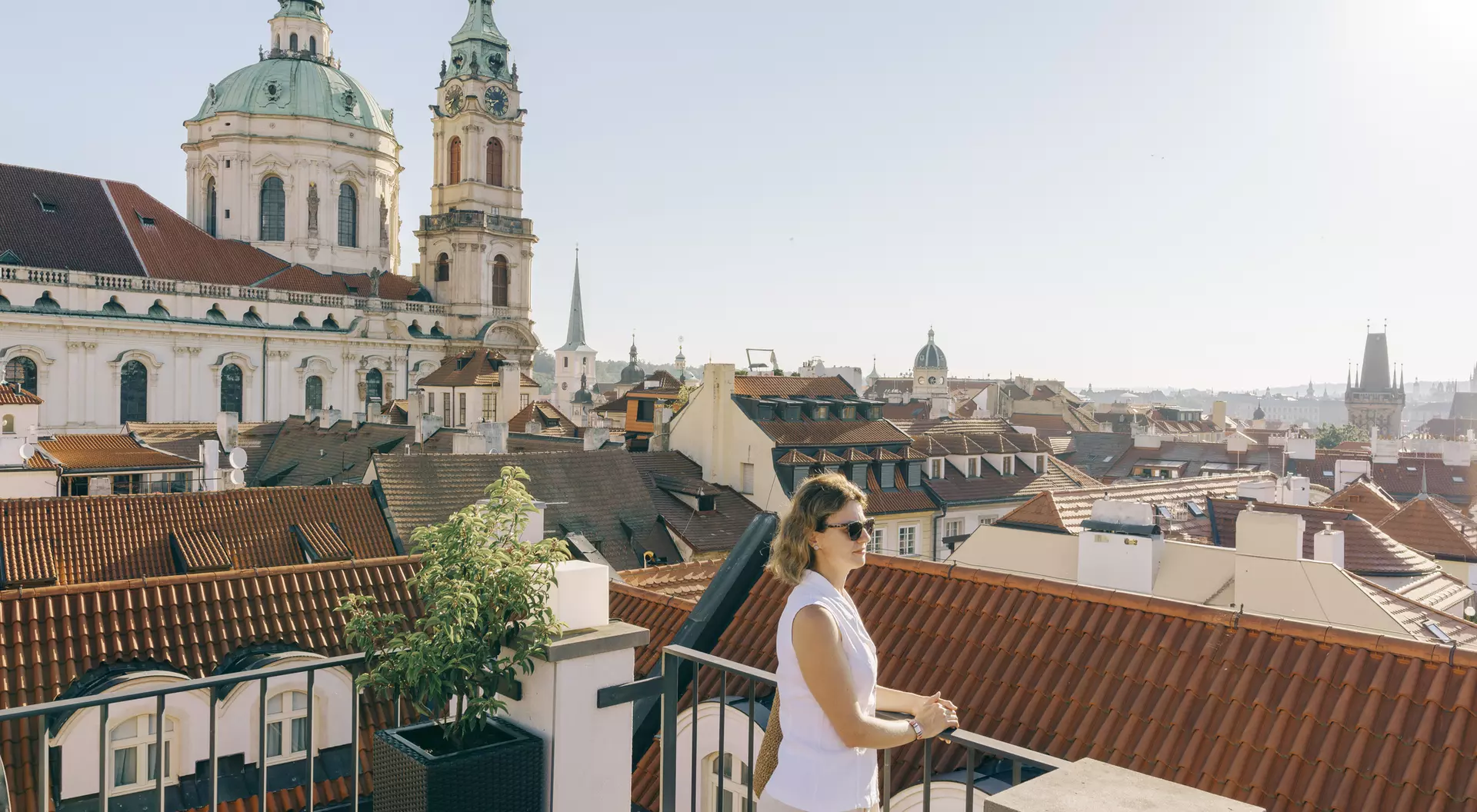 Woman standing on a rooftop terrace overlooking a cityscape with historic buildings and church domes under clear skies.