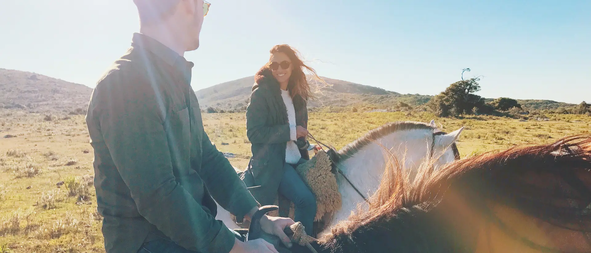 Two people horseback riding in a sunny landscape, smiling and looking at each other.