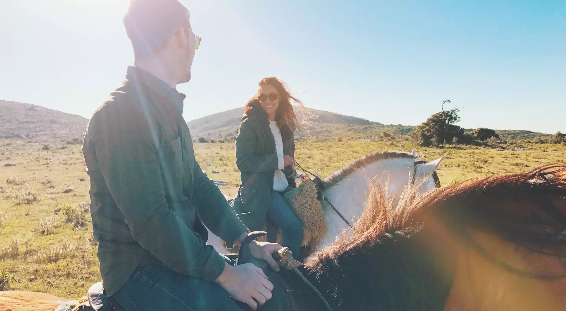 Two people horseback riding in a sunny landscape, smiling and looking at each other.