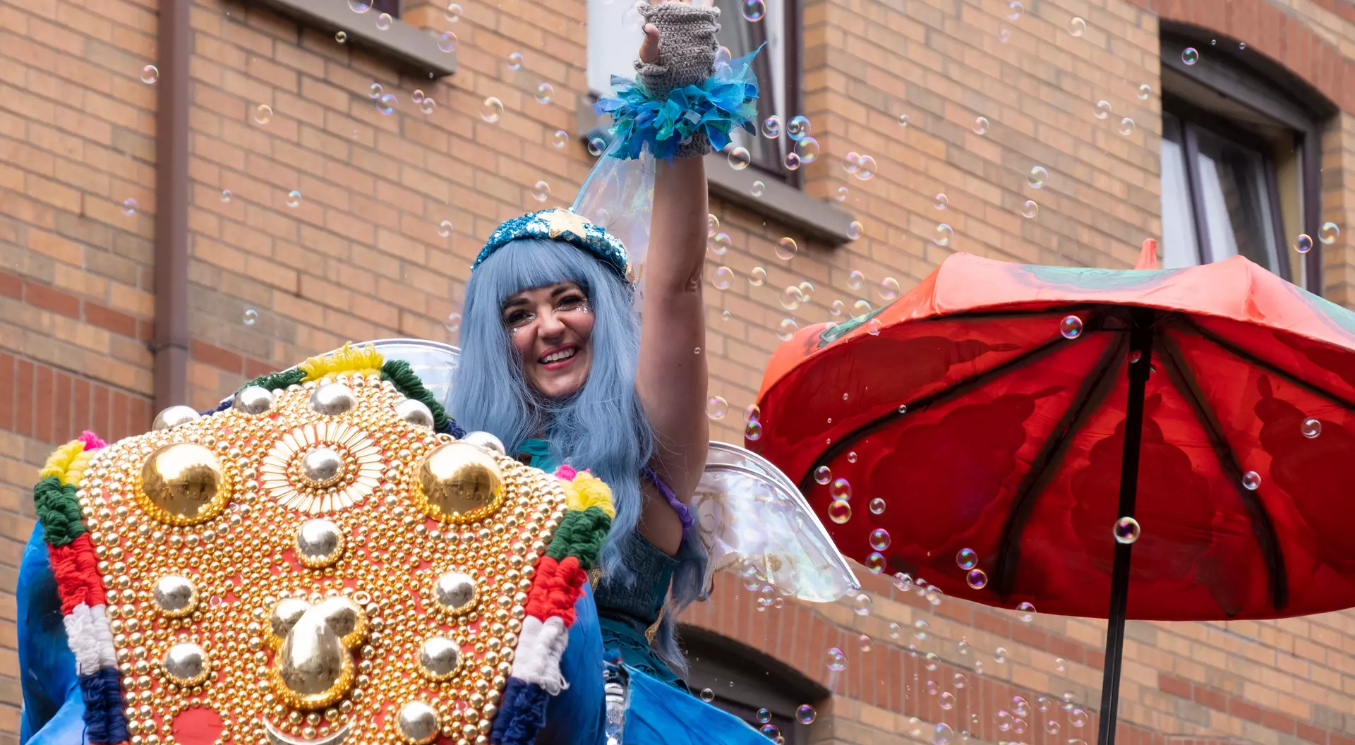 A performer with blue hair waves from a decorated float, surrounded by bubbles and a red umbrella.