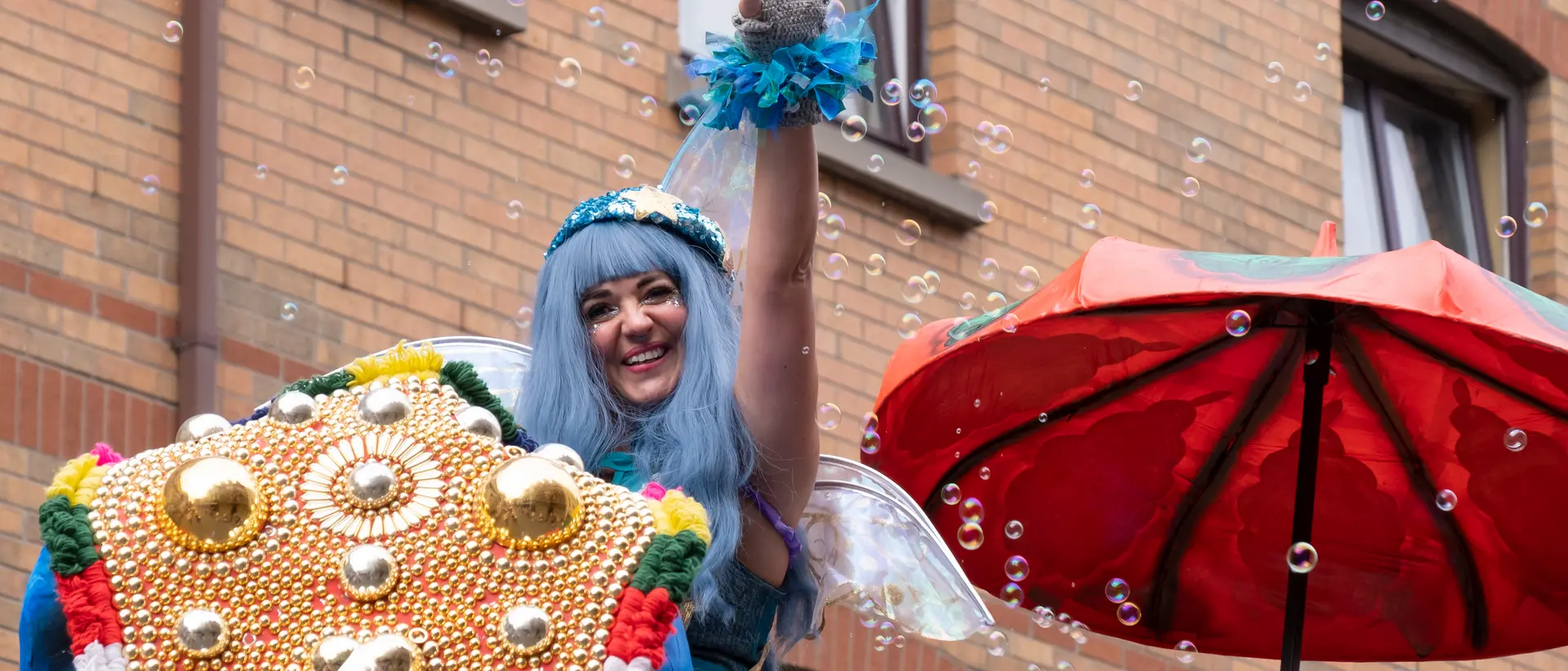 A performer with blue hair waves from a decorated float, surrounded by bubbles and a red umbrella.