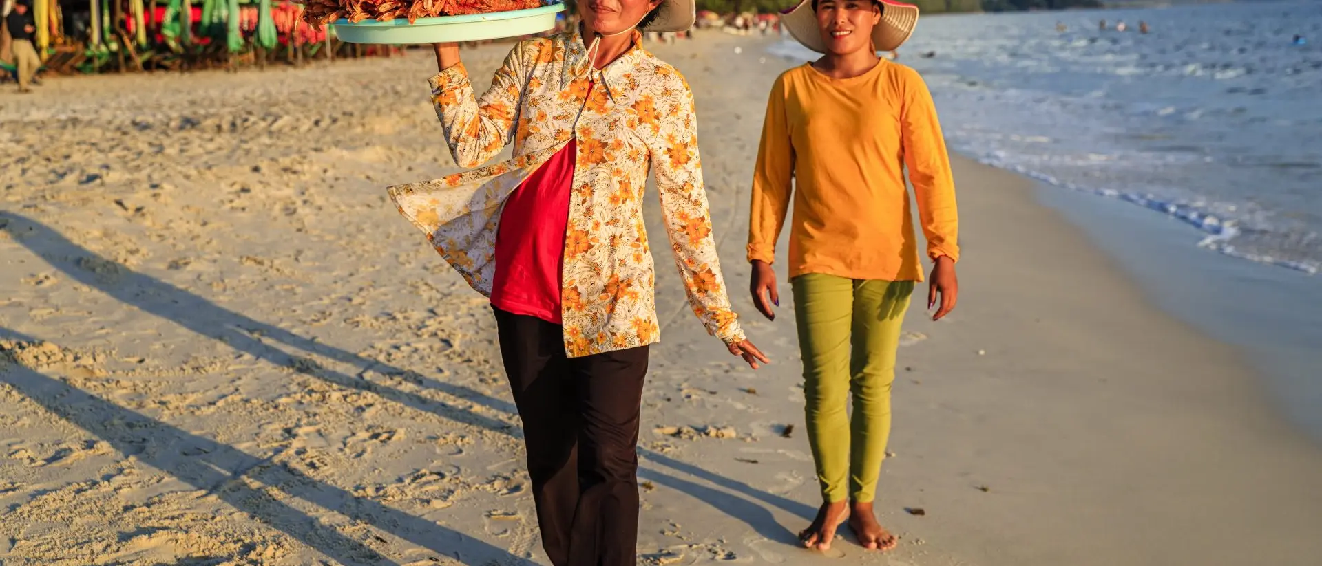Two women walk along a beach, carrying trays of food on their heads, with a calm sea and trees in the background.