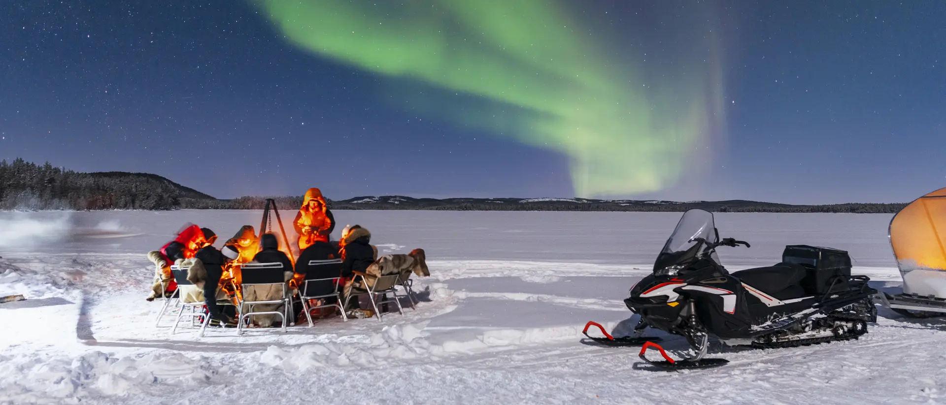 People sit by a fire on snow, watching the Northern Lights above, with a snowmobile parked nearby.