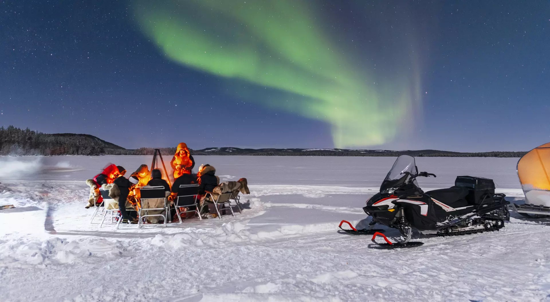 People sit by a fire on snow, watching the Northern Lights above, with a snowmobile parked nearby.