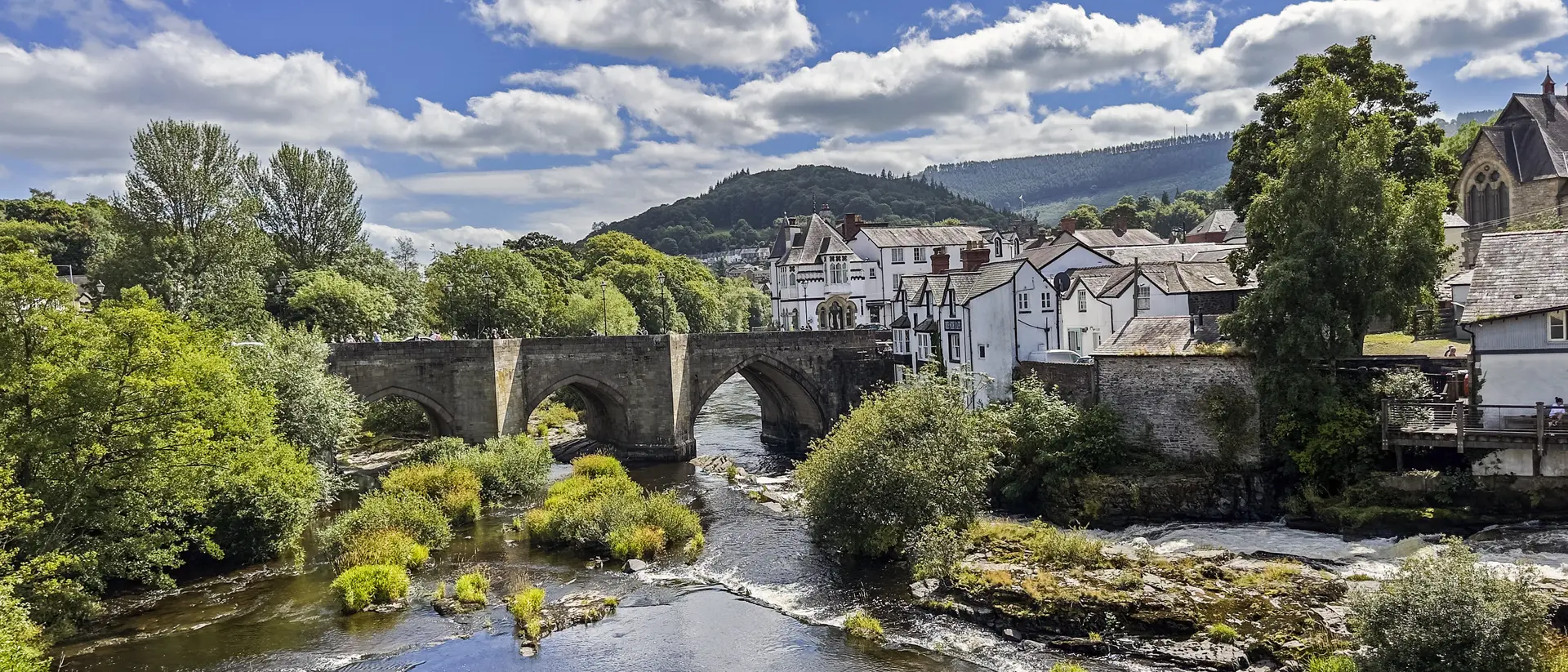 Stone bridge over a river, surrounded by lush greenery and quaint buildings under a partly cloudy sky.