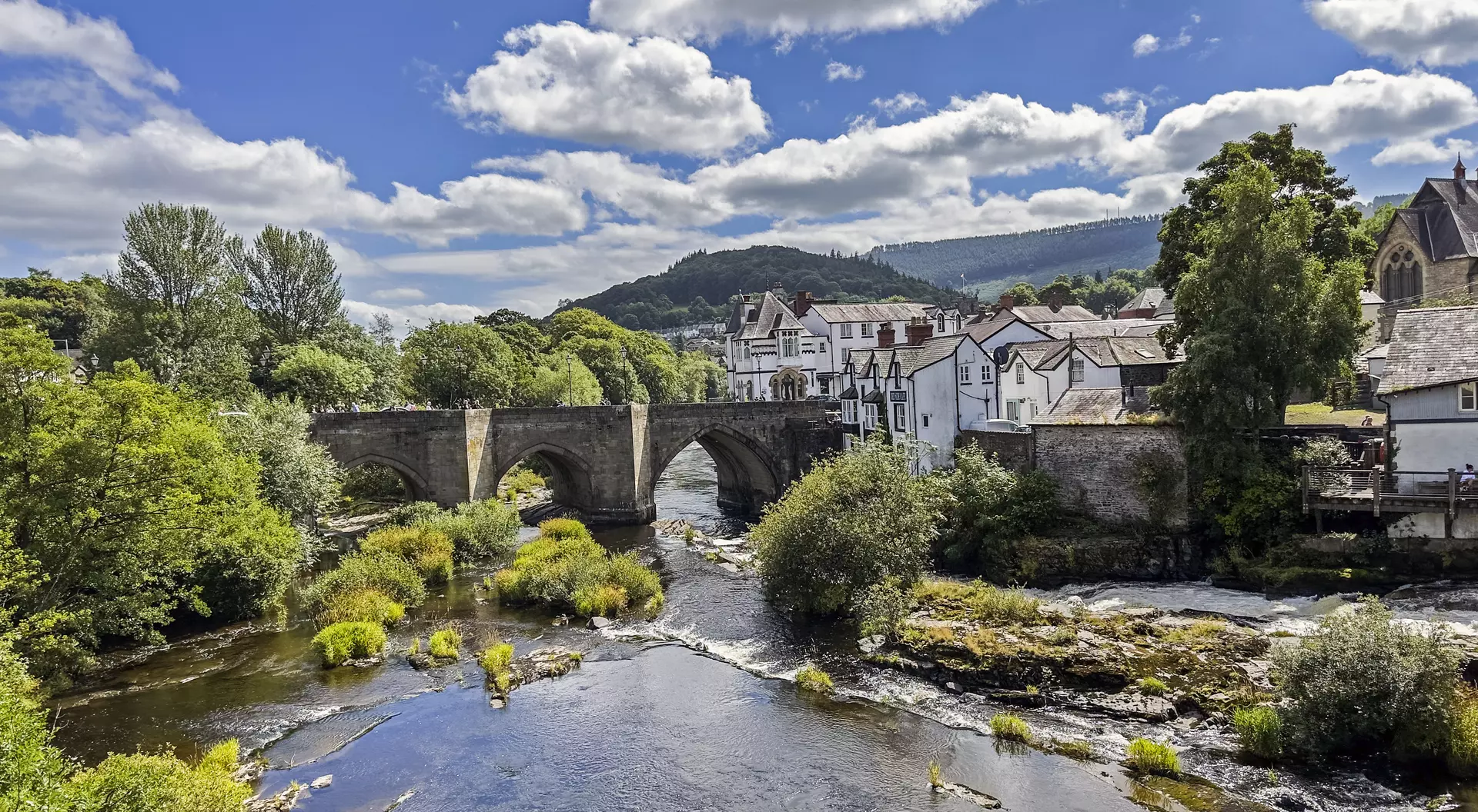 Stone bridge over a river, surrounded by lush greenery and quaint buildings under a partly cloudy sky.