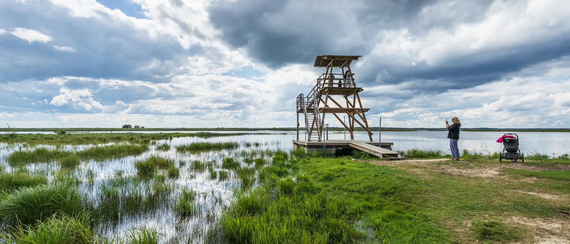 A wooden observation tower by a marsh, with a person taking photos and a stroller nearby under a cloudy sky.