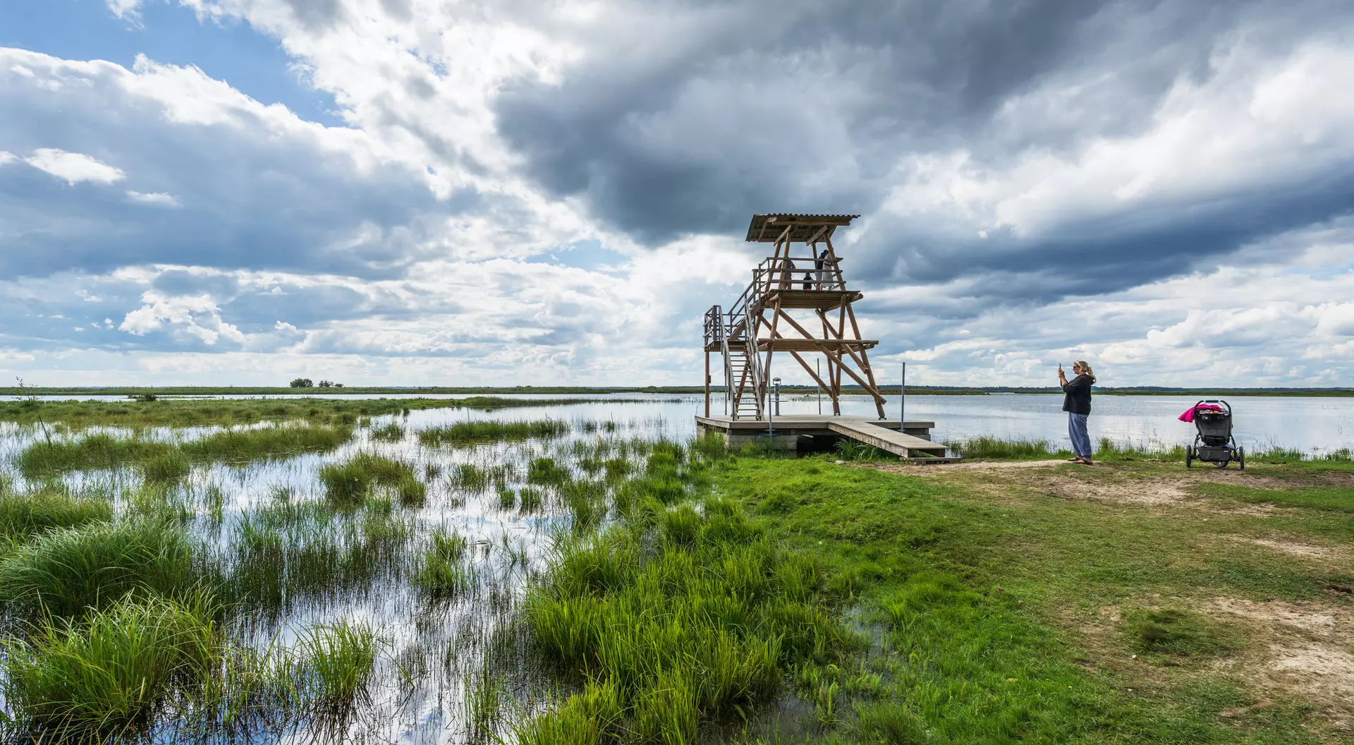 A wooden observation tower by a marsh, with a person taking photos and a stroller nearby under a cloudy sky.
