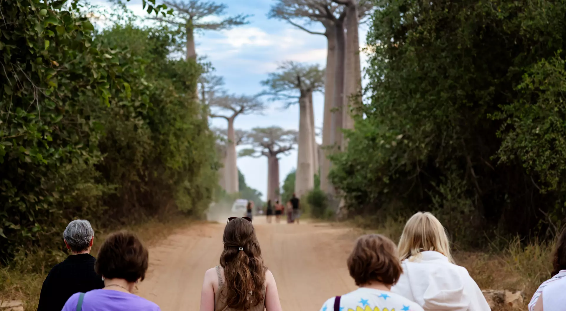 A group of people walks down a dirt path lined with tall baobab trees under a cloudy sky.