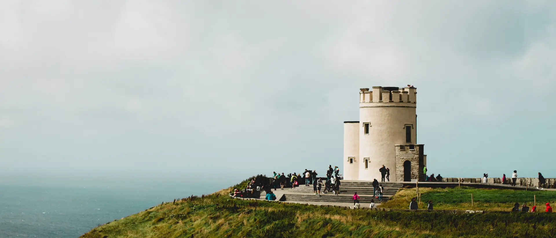 A scenic view of a round white tower on a cliff, surrounded by people, with a backdrop of the sea and cloudy sky.