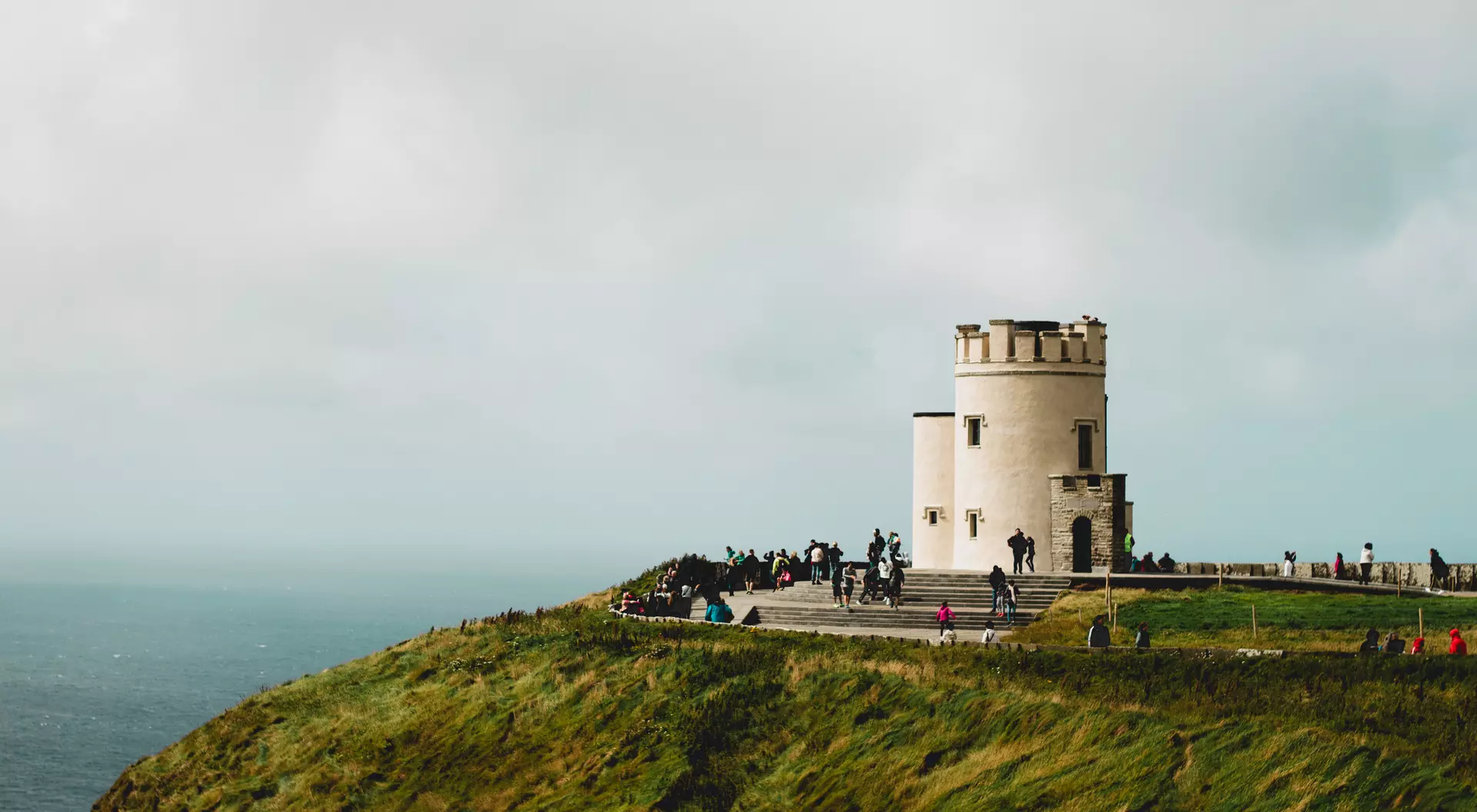 A scenic view of a round white tower on a cliff, surrounded by people, with a backdrop of the sea and cloudy sky.