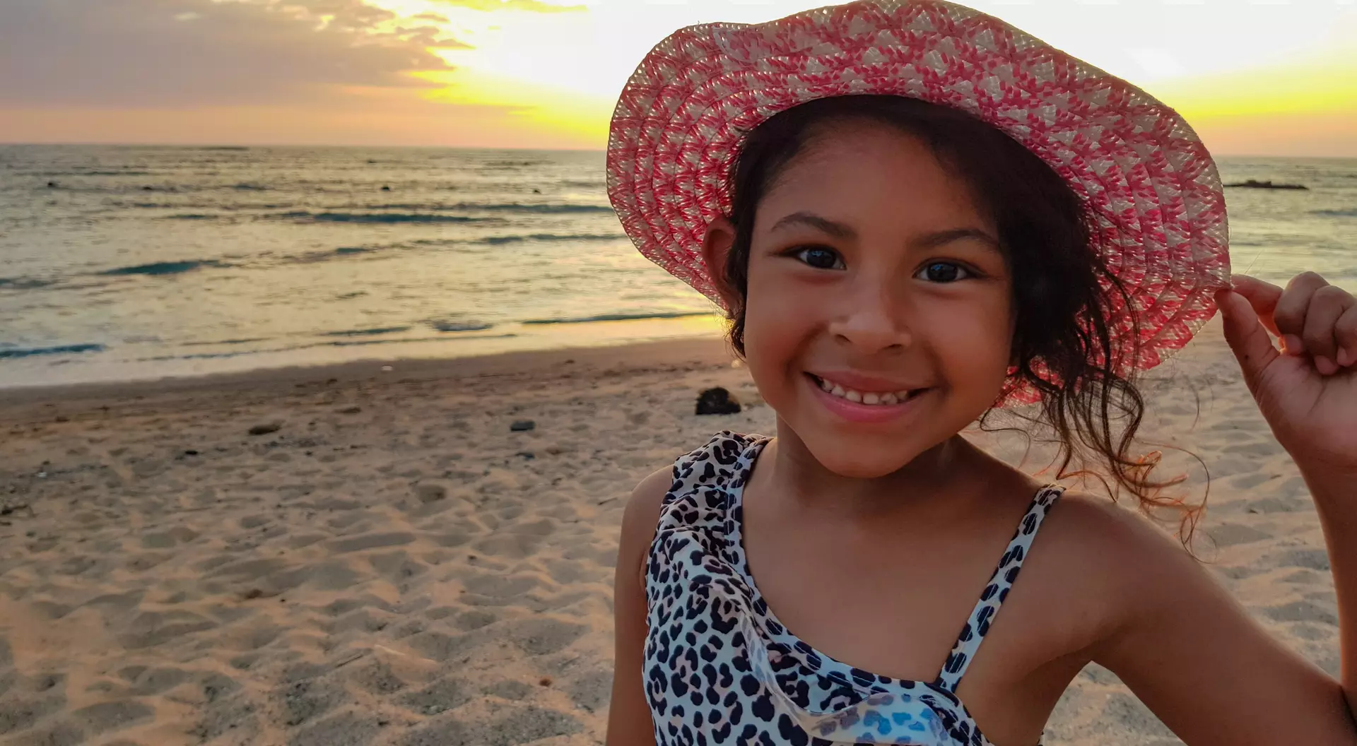 Smiling girl in a leopard print swimsuit and a pink sun hat, standing on the beach at sunset. Ocean waves in the background.