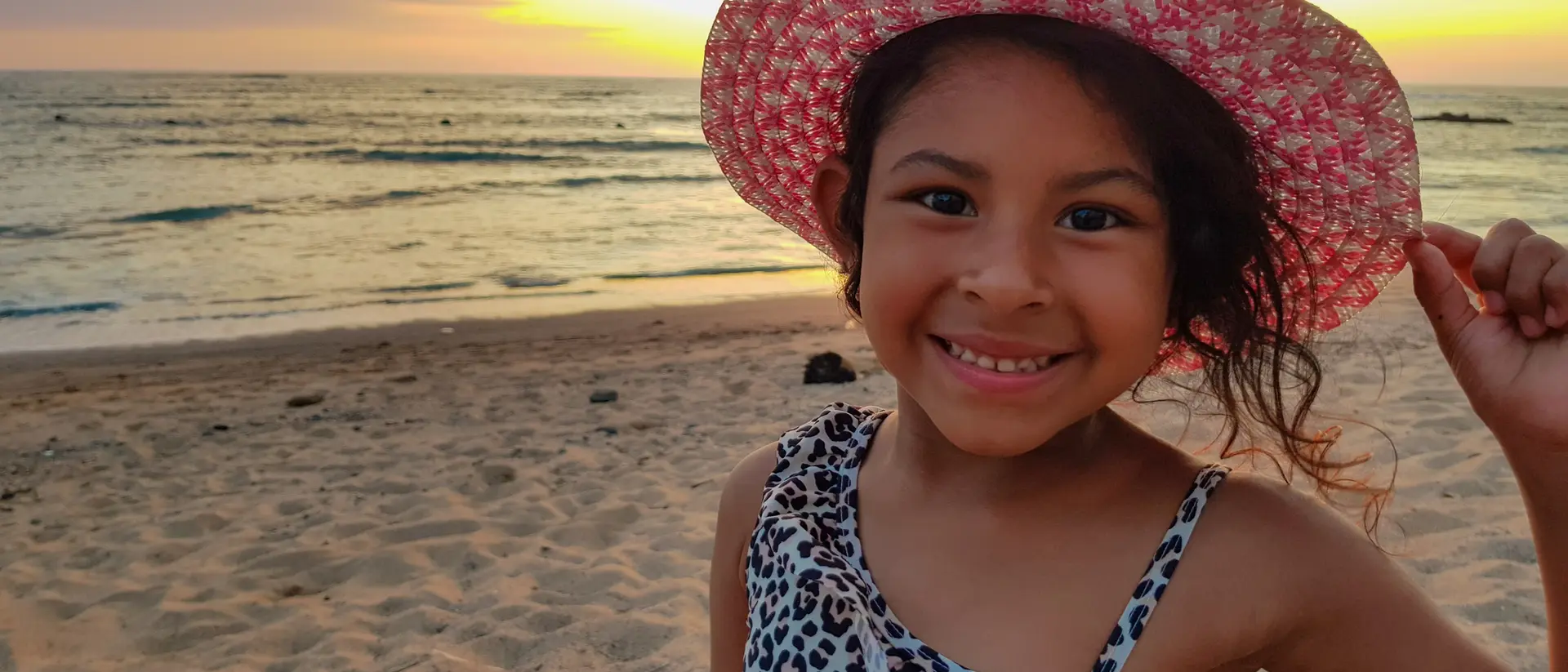 Smiling girl in a leopard print swimsuit and a pink sun hat, standing on the beach at sunset. Ocean waves in the background.